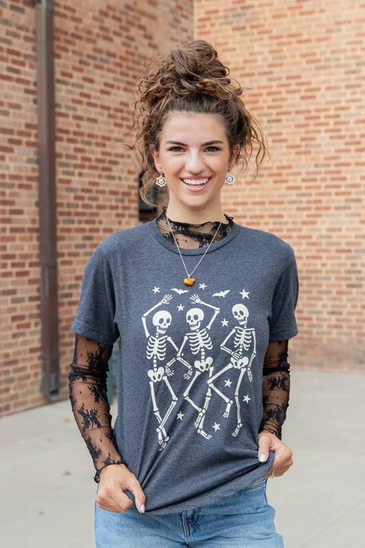 A woman wearing a dark gray graphic t-shirt with a white skeleton print, styled with sheer black lace long sleeves and blue jeans, standing in front of a brick wall.