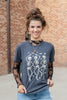A woman wearing a dark gray graphic t-shirt with a white skeleton print, styled with sheer black lace long sleeves and blue jeans, standing in front of a brick wall.