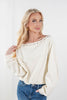 A blonde woman wearing a cream-colored, long-sleeve off-the-shoulder top with silver stud detailing along the neckline, paired with blue jeans, against a plain white background.