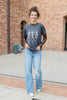 A woman wearing a dark gray graphic t-shirt with a white skeleton print, styled with sheer black lace long sleeves and blue jeans, standing in front of a brick wall.