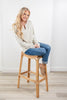 A woman with long blonde hair sits on a wooden bar stool, wearing a cream and gray striped long-sleeve henley top, blue jeans, and tan loafers in a bright room with white walls and light wood flooring.