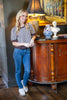 A woman stands indoors next to a wooden cabinet, wearing a black and white gingham short-sleeve top with dark ruffled trim, blue jeans, and white loafers.