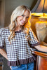 A smiling blonde woman wearing a brown and white gingham short-sleeve top with ruffled trim, paired with blue jeans, posed indoors next to a lit table lamp.