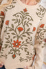 A close-up of a cream-colored knit sweater featuring a large floral and vine pattern in olive green and burnt orange, worn by a woman.