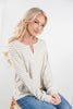 A blonde woman wearing a cream, tan and  gray striped long-sleeve brushed henley shirt with blue jeans, sitting against a plain white studio background.