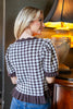 A woman wearing a brown and white gingham short-sleeve top with ruffled trim, shown from the back in an indoor setting with a lamp in the background.