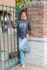 A woman wearing a dark gray graphic t-shirt with a white skeleton print, styled with sheer black lace long sleeves and blue jeans, standing in front of a green fence.