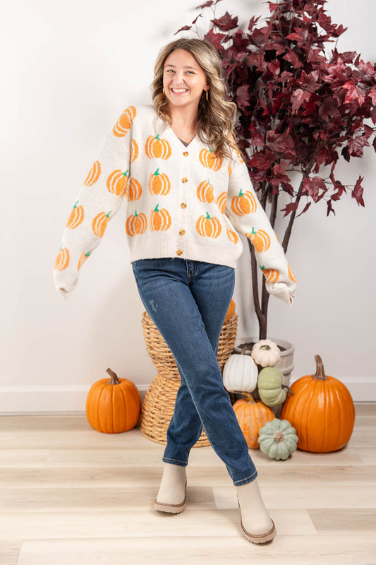 A woman modeling a cream knit cardigan with an orange and green pumpkin pattern, paired with blue jeans and beige boots, standing in a decorated autumn room with pumpkins and a potted plant.