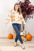 A woman modeling a cream knit cardigan with an orange and green pumpkin pattern, paired with blue jeans and beige boots, standing in a decorated autumn room with pumpkins and a potted plant.