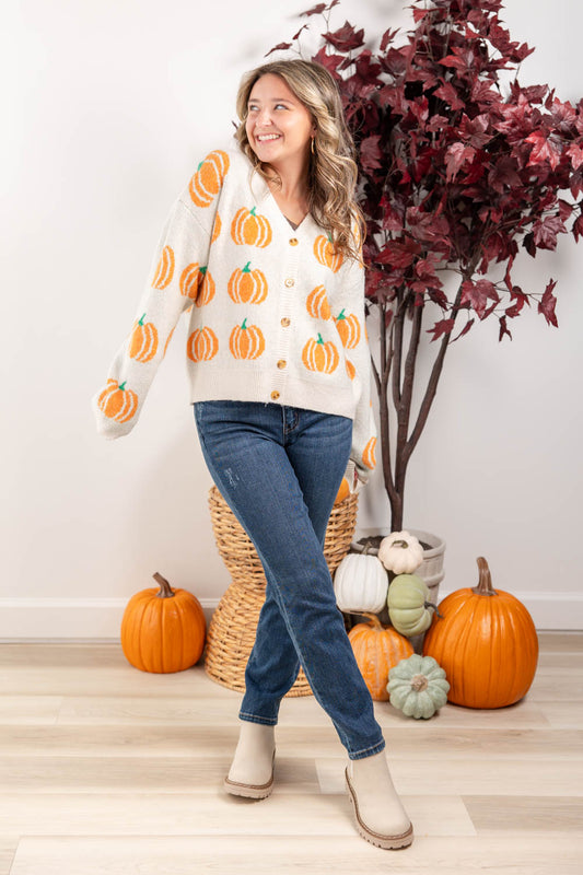 A woman wearing a cream knit cardigan with an orange pumpkin pattern, blue jeans, and beige boots, standing in a fall-themed room decorated with pumpkins and a potted tree.