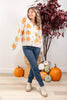 A woman wearing a cream knit cardigan with an orange pumpkin pattern, blue jeans, and beige boots, standing in a fall-themed room decorated with pumpkins and a potted tree.