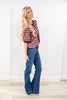 A woman modeling a short-sleeve maroon and pink striped top with blue flare jeans, standing in a bright room with a white wall and light wood floor.