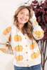 A woman smiling and looking upward while wearing a cream knit cardigan with an orange pumpkin pattern and gold buttons, paired with blue jeans. She is standing indoors against a light wall with dark red foliage in the background.