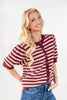 A smiling blonde woman wearing a short-sleeve knit cardigan with horizontal burgundy and pink stripes, paired with blue jeans, against a plain white studio background.