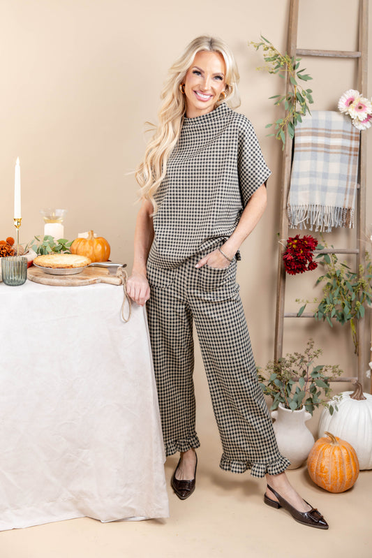 A woman modeling a matching two-piece houndstooth jumpsuit set in a cozy, autumn-themed indoor setting with pumpkins, a table, and a ladder backdrop.