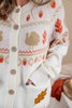 A close-up of a person wearing a cream knit cardigan with autumn-themed patterns including a turkey, acorns, and orange leaves. The cardigan features a button-front closure and a front pocket with an embroidered leaf detail.