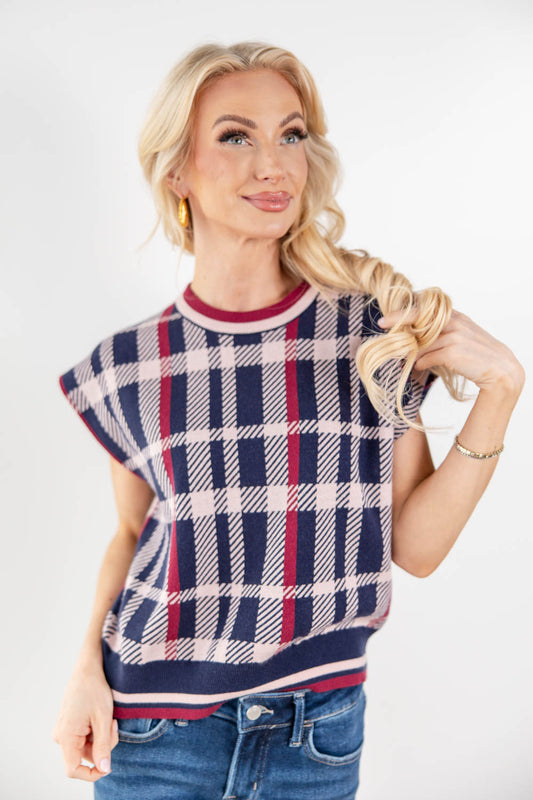 A blonde woman wearing a cap-sleeve knit sweater with a navy, pink, and white plaid pattern, paired with blue jeans, against a plain white studio background.