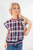 A blonde woman wearing a cap-sleeve knit sweater with a navy, pink, and white plaid pattern, paired with blue jeans, against a plain white studio background.