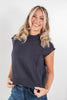 A smiling blonde woman wearing a dark charcoal gray ribbed knit mock-neck sweater with cap sleeves and blue jeans, standing against a light gray studio background.