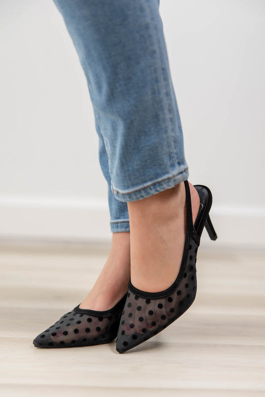 A women wearing slingback high heels featuring a black leather upper with mesh black polka dots, set against a white wall and wood floor.