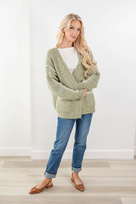 A woman modeling an oversized sage green knit cardigan with white blanket stitch sleeve details, paired with blue jeans and brown flats in a bright indoor setting.