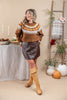 A woman modeling an autumn-themed outfit in a styled indoor studio setting. She is wearing a brown knit sweater with a cream and orange Nordic-style collar pattern, a dark brown faux-leather mini skirt, and tan knee-high leather boots.