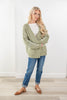 A woman modeling an oversized sage green knit cardigan with white blanket stitch sleeve details, paired with blue jeans and brown flats in a bright indoor setting.