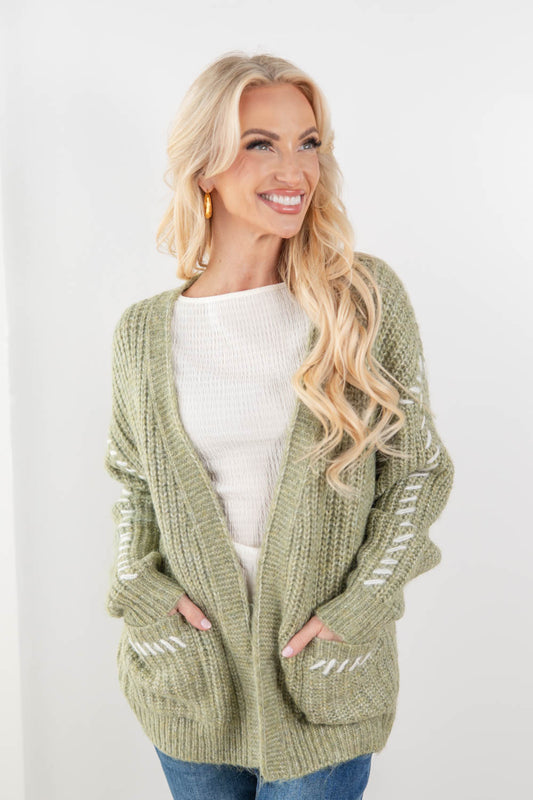 A smiling blonde woman wearing a chunky sage green open-front cardigan with white embroidered stripe details on the sleeves and pockets, layered over a white ribbed top and blue jeans, against a plain white studio background.