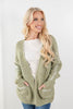A smiling blonde woman wearing a chunky sage green open-front cardigan with white embroidered stripe details on the sleeves and pockets, layered over a white ribbed top and blue jeans, against a plain white studio background.