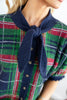 A close-up of a woman wearing a green, red, and navy plaid knit top with a contrasting navy blue knotted collar.