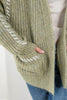 Close-up of a person wearing a chunky knit cardigan in a muted sage green color, featuring white contrast knit details along the sleeve seams and a pocket with a hand inserted.