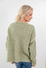 A woman with blonde hair is shown from the back wearing a chunky knit sage green sweater and blue jeans against a plain white background.