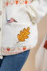 Close-up of a cream knit cardigan with a large embroidered gold oak leaf on the pocket, featuring orange and beige autumn-themed patterns along the trim, worn with blue jeans.