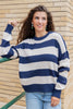 A smiling woman wearing a navy and off-white horizontally striped knit sweater, paired with jeans, standing against a brick wall.