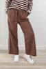 A person wearing brown wide-leg drawstring pants with a white lace stripe detail down the side, paired with a brown and white striped long-sleeve top and white sneakers, standing against a light gray wall and wood floor.