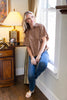 A woman wearing a brown short-sleeve button-up shirt with white polka dots and blue jeans, leaning against a white window frame in a warmly lit room.