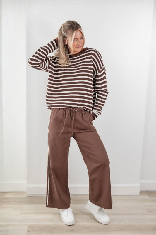 A woman modeling a brown and white horizontally striped long-sleeve knit sweater with wide-leg brown pants with lace trim and white sneakers, standing in a bright room with white walls and light wood flooring.