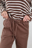 A close-up of a person wearing brown drawstring sweatpants and a brown-and-white striped knit sweater, with both hands pulling the waistband of the pants.