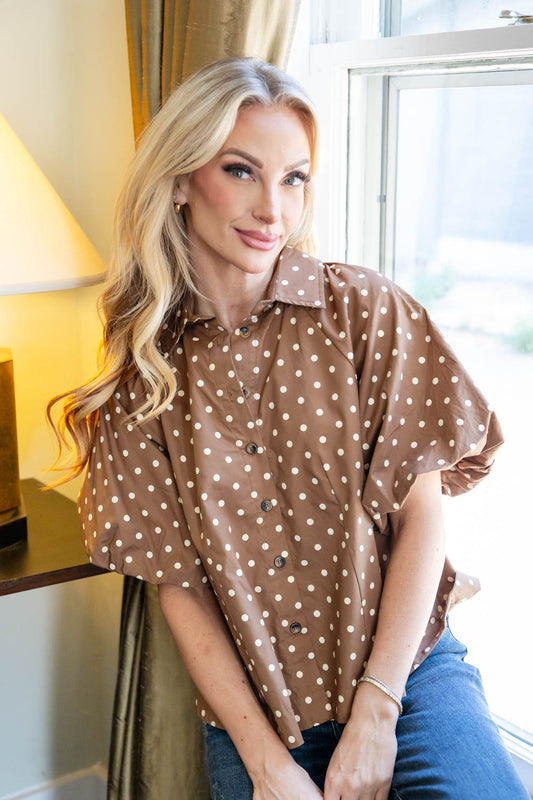 A woman with blonde hair wearing a brown short-sleeve button-up shirt with white polka dots and blue jeans, sitting in a brightly lit room.