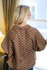 A woman with blonde hair is shown from behind wearing a brown short-sleeve blouse with white polka dots, standing in front of a window.
