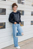 A woman stands against a white siding wall wearing a black crewneck sweatshirt with a gray 'BOO' graphic, light-wash distressed jeans, and white sneakers.