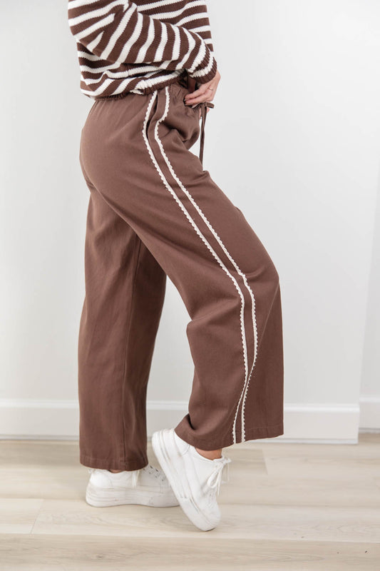 A person wearing brown wide-leg pants with white lace decorative side stripes, paired with a brown-and-white striped top and white sneakers, standing indoors against a light wall.