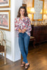 A woman stands in a warmly lit room wearing a long-sleeve button-up blouse with a red, pink, and white floral pattern, paired with blue skinny jeans and brown slide sandals.