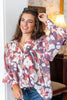 A woman wearing a long-sleeve button-up blouse with a large floral print in dusty pink, red, white, and purple, paired with blue jeans, standing in a warmly lit indoor setting.
