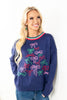 A woman wearing a navy blue crewneck sweater decorated with colorful embroidered bows in pink, green, and red, paired with blue jeans.