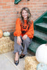 A woman sits on outdoor steps wearing a bright orange crewneck sweatshirt with a black jack-o'-lantern graphic sweater, paired with black and white gingham ruffled pants and black pointed flats with gold hardware. She is posed on straw bales with pumpkins against a brick wall.