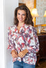 A woman wearing a long-sleeve button-up blouse with a floral pattern in pink, red, and white over a sheer mauve-purple base, paired with blue jeans, standing in a warmly lit indoor setting.