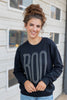 A woman with her hair in a messy bun smiles while wearing a black crewneck sweatshirt with a large gray 'BOO' graphic on the front, paired with light blue jeans, standing in front of a white house exterior.