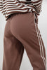Close-up of a woman wearing brown pants with white lace side stripes and a brown-and-white striped top, shown from the waist down against a light gray background.