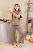 A woman modeling a brown and beige gingham matching lounge set, featuring a short-sleeve top and wide-leg pants with ruffled hems, posed in a cozy autumn-themed studio setting.